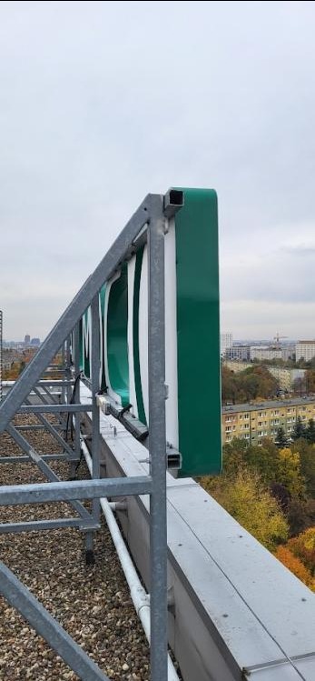 Wysoka konstrukcja reklamowa z zielonymi elementami na dachu budynku, widok na miasto w tle, pochmurne niebo. Metalowa rama wspierająca reklamę.