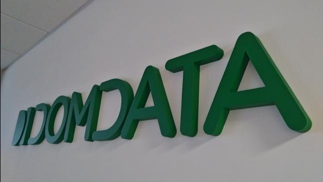 Zielone, przestrzenne litery 'IDOMDATA' na białej ścianie, wykonane w Poznaniu. Litery tworzą cień, co dodaje im trójwymiarowości. Ujęcie z dołu, lekko z boku.