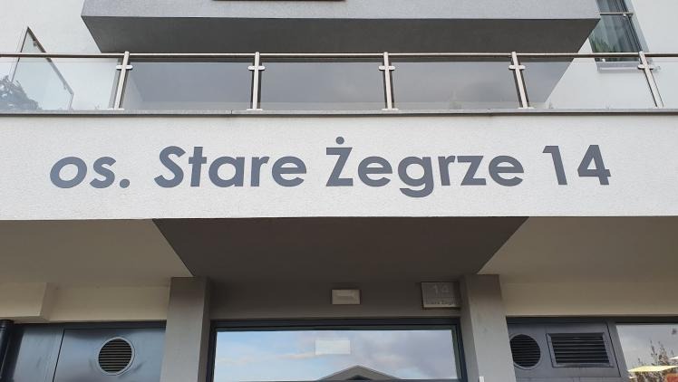 Szary napis 'os. Stare Żegrze 14' na elewacji budynku, nad wejściem. Widoczny balkon z szklaną balustradą i fragmenty okien. Ujęcie z dołu.