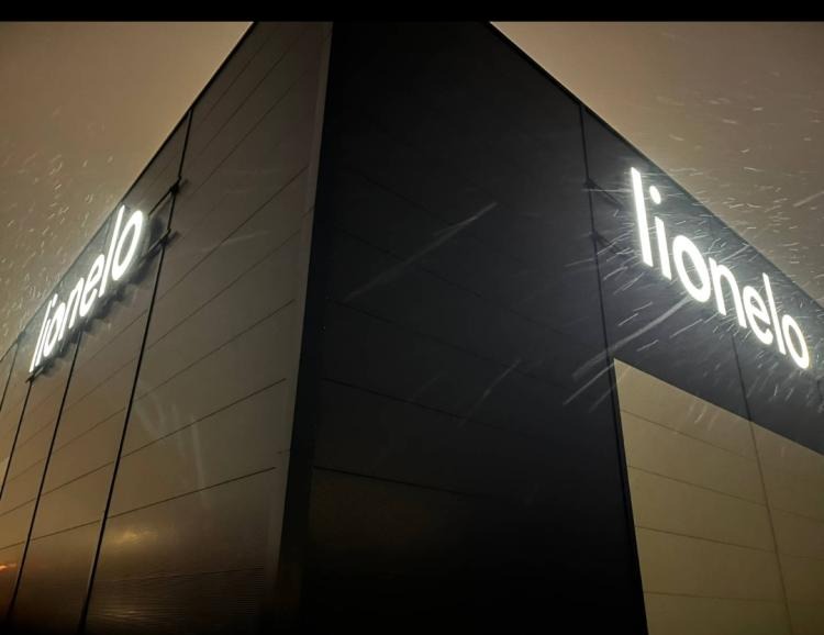 Elewacja budynku z podświetlanym neonowym napisem 'lionelo' w zimowej scenerii. Widok z dołu, padający śnieg. Nocne oświetlenie.