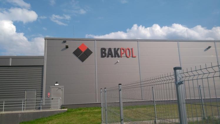 Szary budynek magazynowy z logo i nazwą firmy BAKPOL w kolorach czerni i czerwieni, widoczne zza metalowego ogrodzenia na tle błękitnego nieba z chmurami.
