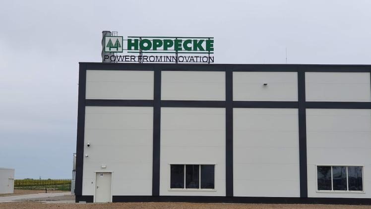 Budynek przemysłowy z logo Hoppecke na dachu, białe ściany z ciemnymi pasami, okna i drzwi. Widok z zewnątrz, pochmurne niebo w tle.