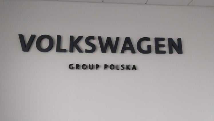 Elegancki napis przestrzenny 'VOLKSWAGEN GROUP POLSKA' w ciemnoszarym kolorze, umieszczony na gładkiej, białej ścianie. Minimalistyczny design, równomierne oświetlenie.