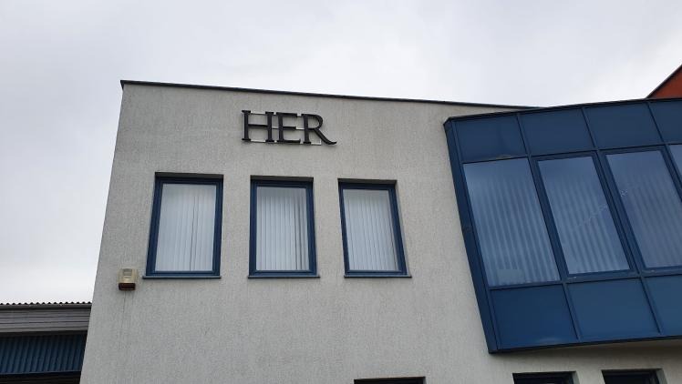 Elewacja budynku z trzema oknami i napisem 'HER' nad nimi. Widoczne okna z białymi żaluzjami i fragment przeszklonej konstrukcji po prawej stronie.