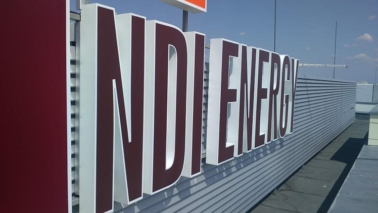 Przestrzenne litery z nazwą firmy 'NDI ENERGY' w kolorze bordowym z białym obramowaniem, zamontowane na szarej, metalowej elewacji budynku. Widok z dołu, błękitne niebo w tle.