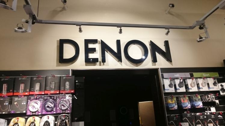 Elegancki szyld z czarnymi, błyszczącymi literami 'DENON' nad wejściem do sklepu, oświetlony reflektorami. Półki z produktami po bokach.