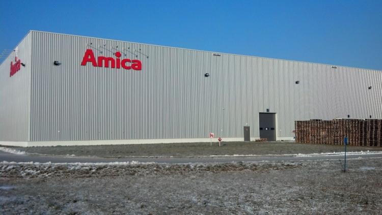 Budynek magazynowy z blachy falistej z czerwonym logo Amica. Widoczne palety i fragment ośnieżonego terenu. Błękitne niebo w tle. Ujęcie z daleka, lekko z dołu.