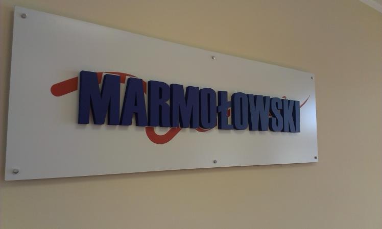 Tablica z nazwą firmy 'MARMOŁOWSKI' wykonana z trójwymiarowych, ciemnoniebieskich liter na białym tle z czerwonym elementem graficznym, zamocowana na ścianie.