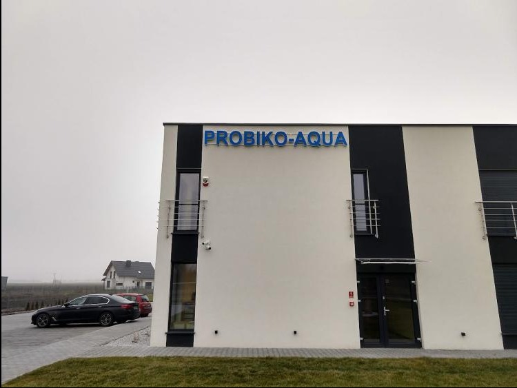 Budynek z niebieskim napisem 'PROBIKO-AQUA' nad wejściem, widoczny samochód i fragment krajobrazu w tle, pochmurny dzień.