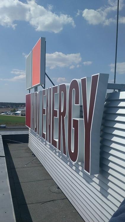 Duży, przestrzenny szyld z napisem 'IND ENERGY' na dachu budynku, z elementami w kolorze czerwonym i szarym, na tle błękitnego nieba z chmurami.