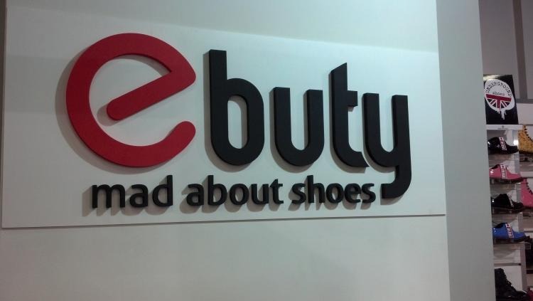 Logo 'e-buty mad about shoes' z czerwonym 'e' i czarnym napisem, umieszczone na białej ścianie w sklepie obuwniczym. W tle widoczne półki z butami.