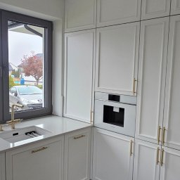 BOX SPÓŁKA Z OGRANICZONĄ ODPOWIEDZIALNOŚCIĄ - Biała zabudowa kuchenna z piekarnikiem i oknem w tle, złote uchwyty, widok na domy w Krakowie. Klasyczny styl, eleganckie wykończenie.