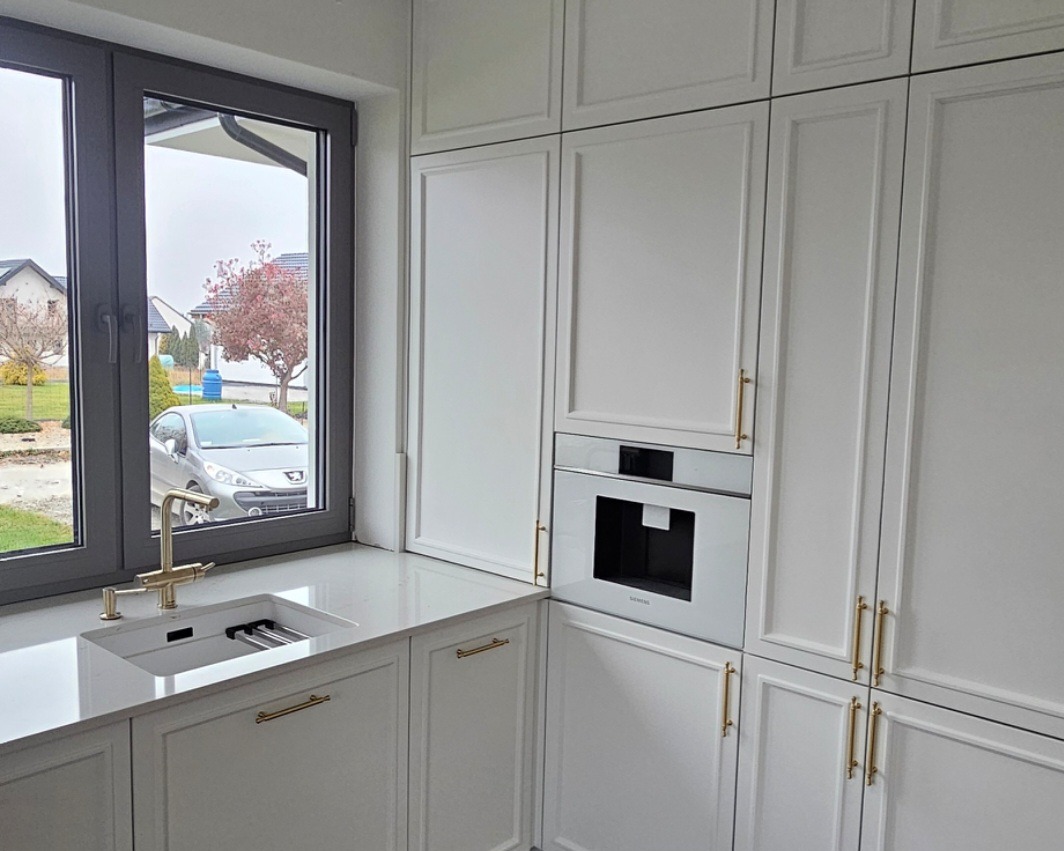 Biała zabudowa kuchenna z piekarnikiem i oknem w tle, złote uchwyty, widok na domy w Krakowie. Klasyczny styl, eleganckie wykończenie.