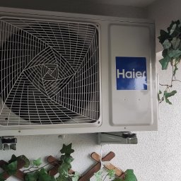 ICE-MAX - Biała jednostka zewnętrzna klimatyzacji Haier zamontowana na balkonie, z dekoracyjną zielenią. Widok na zewnątrz przez balustradę.