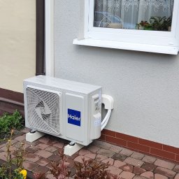 ICE-MAX - Zewnętrzna jednostka klimatyzacji Haier zamontowana na kostce brukowej przy ścianie domu z oknem. Widoczne rośliny w tle i rynna spustowa.
