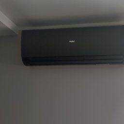 ICE-MAX - Czarny klimatyzator Haier zamontowany na szarej ścianie w nowoczesnym wnętrzu, widok z okna na zewnątrz. Minimalistyczny design.