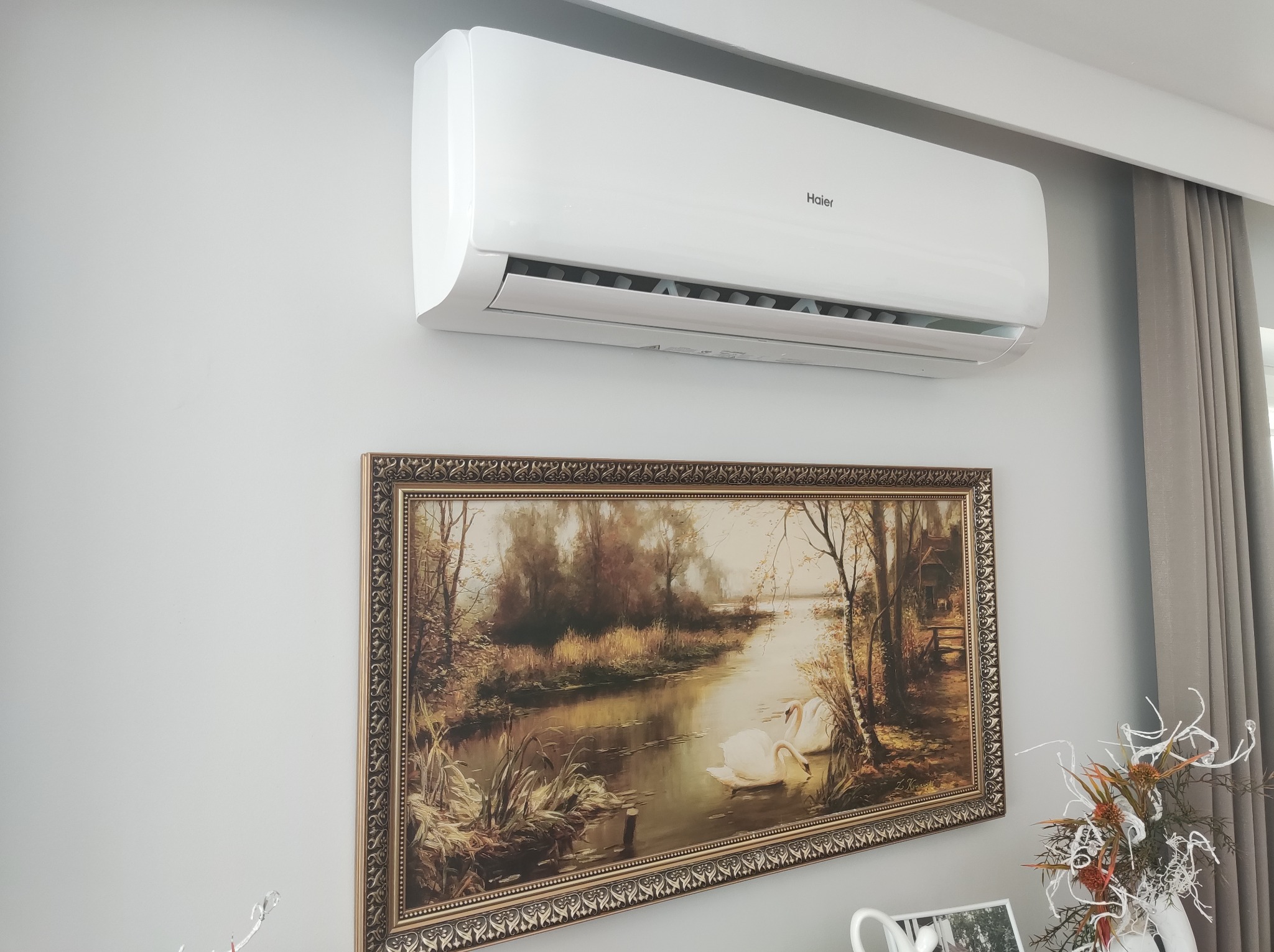 Biały klimatyzator Haier na ścianie nad obrazem z łabędziami w złotej ramie. Szare zasłony w tle. Widok z dołu, oświetlenie dzienne.