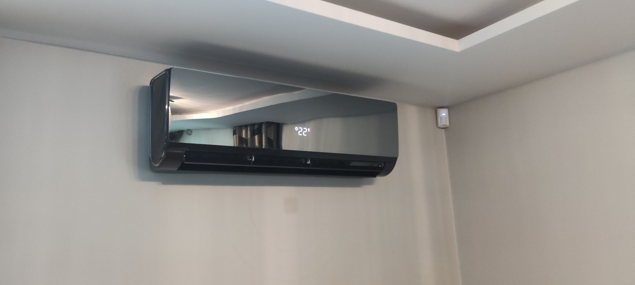 Czarny klimatyzator ścienny z wyświetlaczem temperatury 22°C, zamontowany w rogu jasnego pomieszczenia z widocznym fragmentem sufitu podwieszanego.