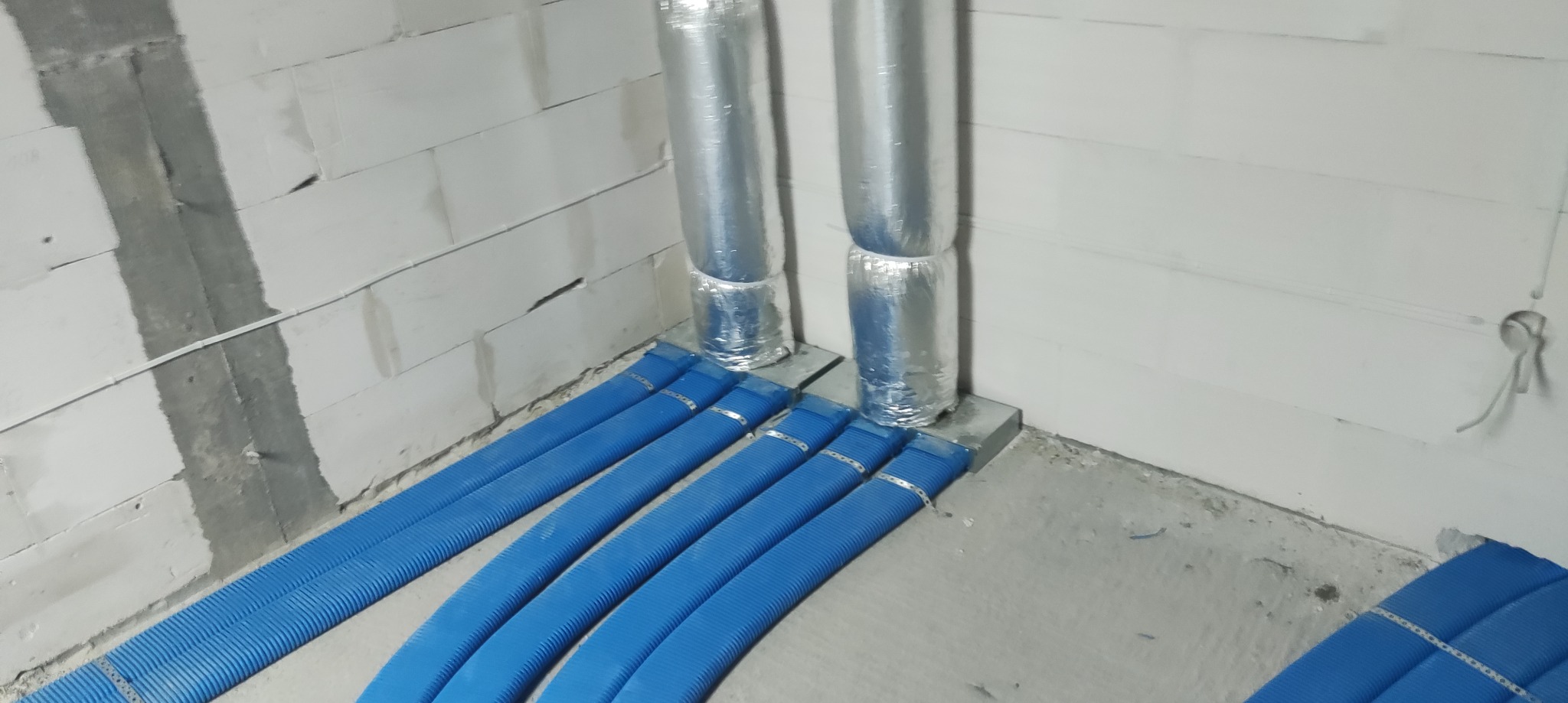 Niebieskie rury wentylacyjne ułożone równolegle na podłodze w surowym wnętrzu, z widocznymi srebrnymi przewodami pionowymi i fragmentem ściany z betonu komórkowego.