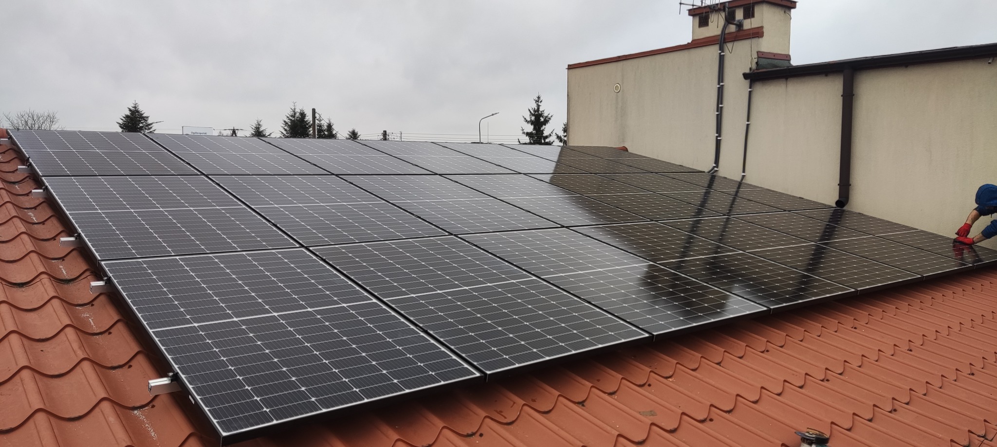 Montaż paneli fotowoltaicznych na dachu z czerwonej dachówki. Widoczny fragment budynku i osoba w trakcie instalacji. Pochmurne niebo w tle.