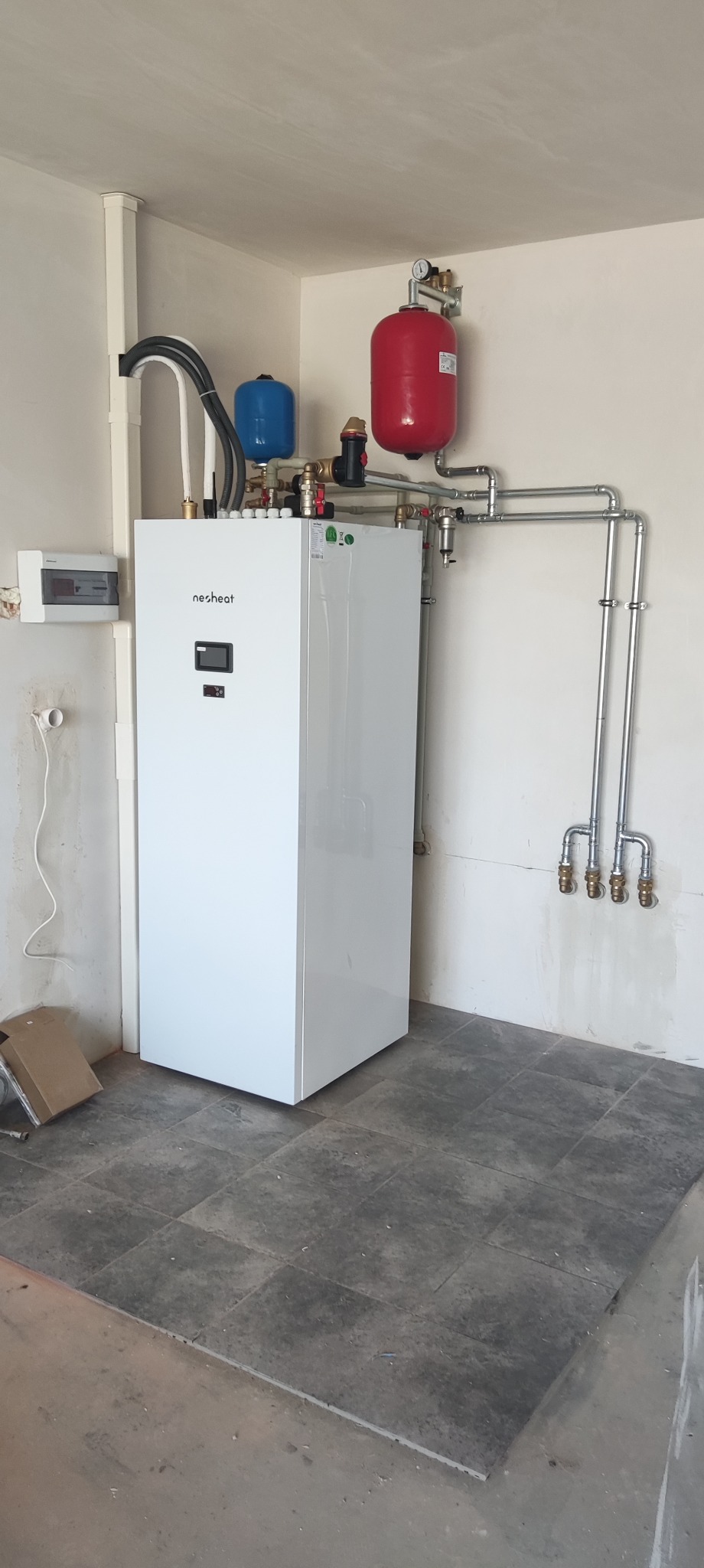 Biała pompa ciepła Nesheat z czerwonym i niebieskim zbiornikiem wyrównawczym, zamontowana w pomieszczeniu z szarą podłogą i instalacją rurową.