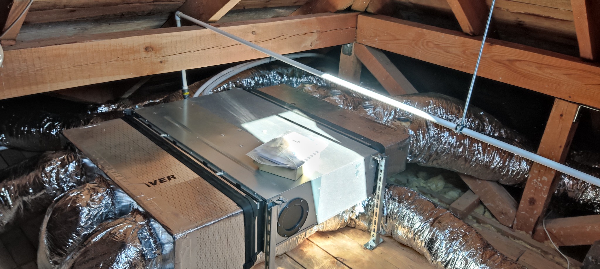 Rekuperator w przestrzeni poddasza, widoczne przewody wentylacyjne w srebrnej izolacji, drewniana konstrukcja dachu i biała rura instalacyjna.