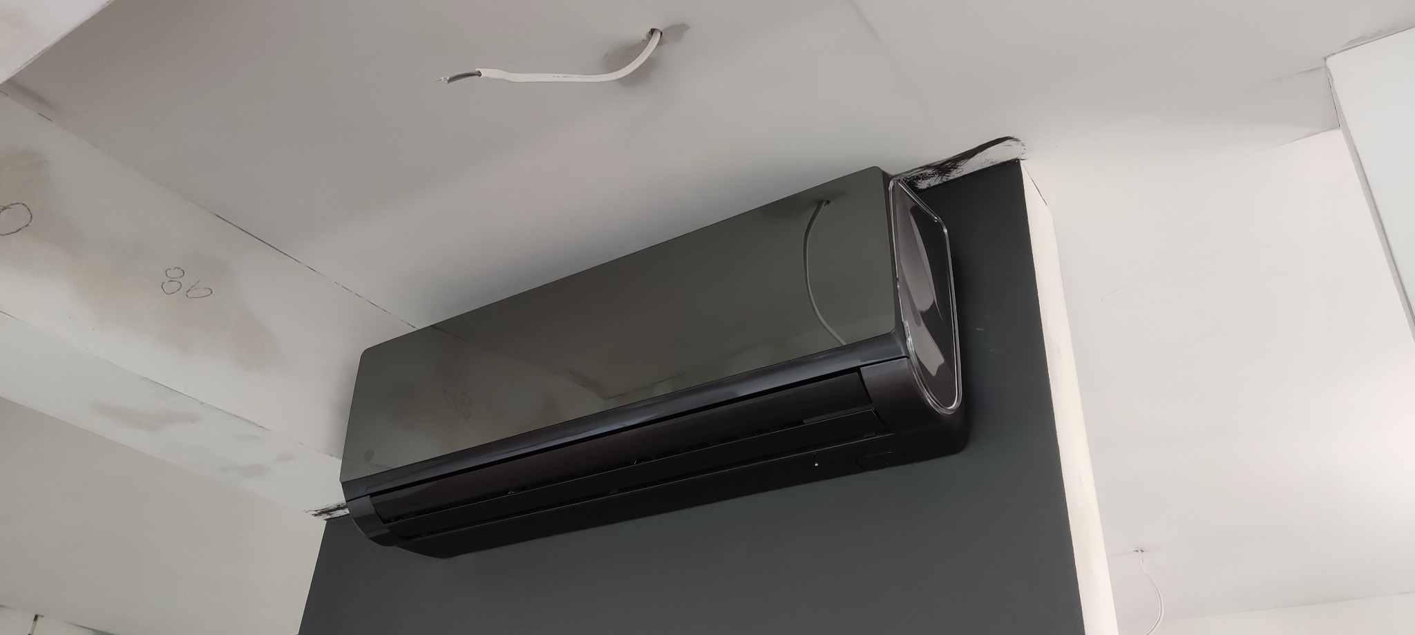 Czarny klimatyzator ścienny zamontowany w pomieszczeniu w trakcie wykańczania, widoczne przewody elektryczne i ślady po malowaniu na suficie.
