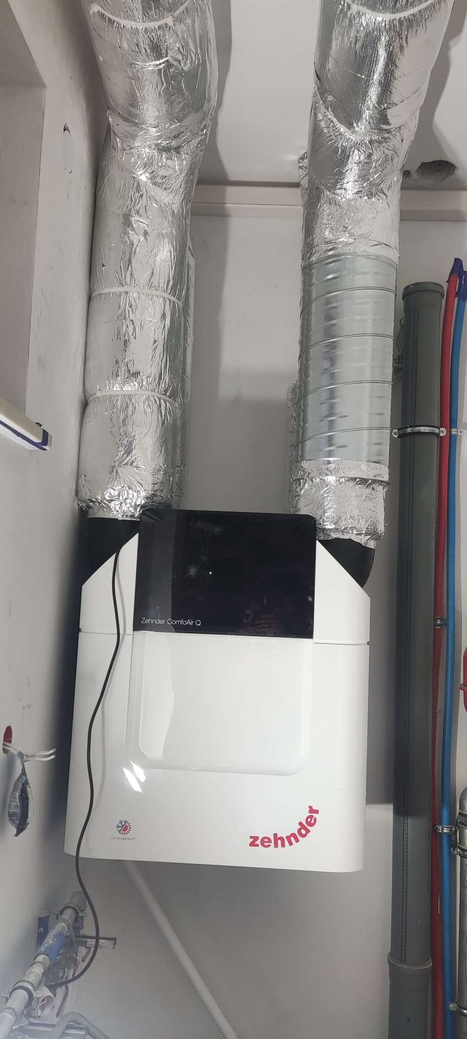 Zamontowana centrala rekuperacyjna Zehnder ComfoAir Q z izolowanymi przewodami wentylacyjnymi w pomieszczeniu technicznym. Widoczne rury instalacyjne.