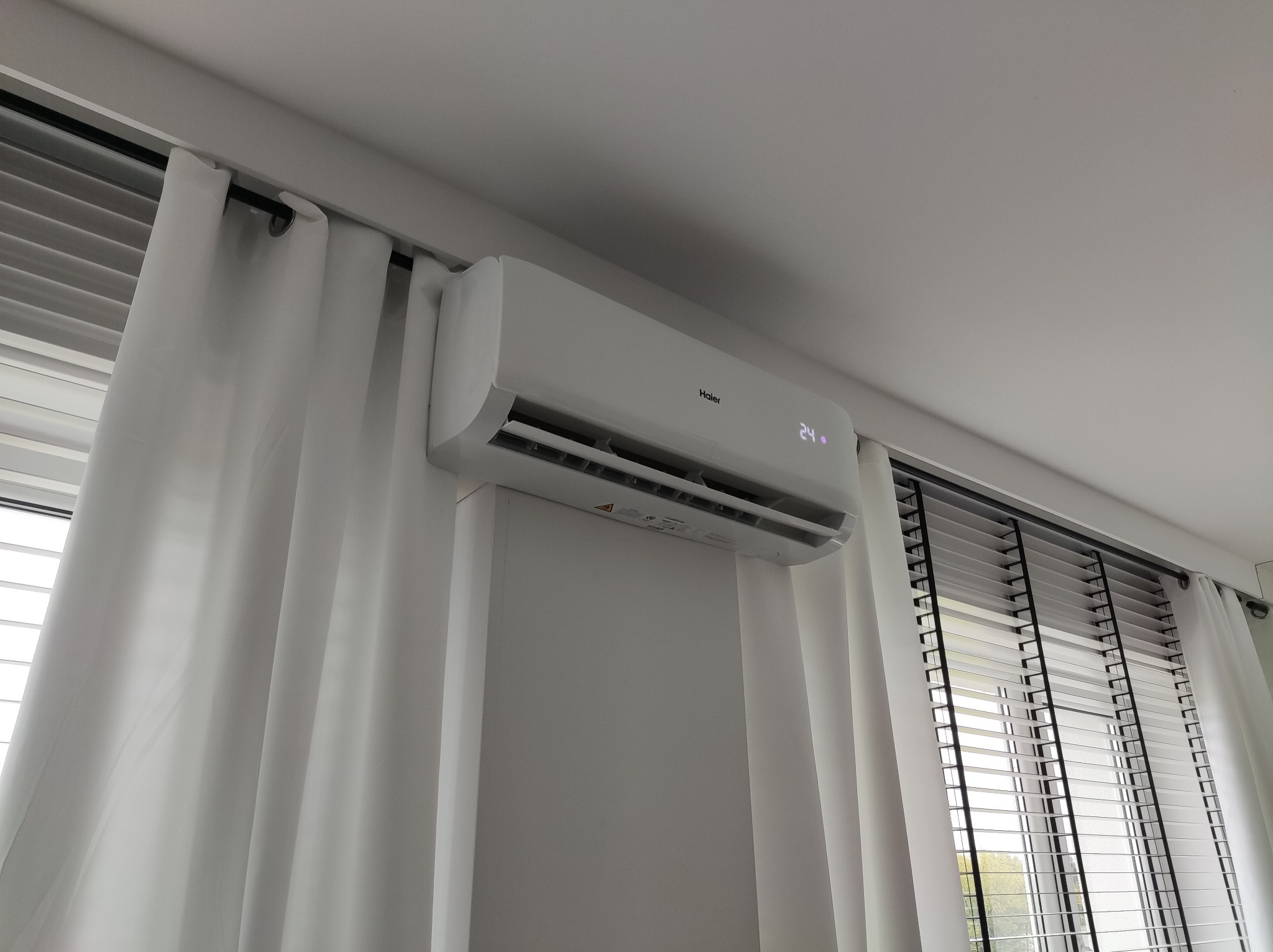 Biały klimatyzator Haier na ścianie, widoczny wyświetlacz z temperaturą 24 stopnie, obok okno z żaluzjami i białe zasłony. Ujęcie z dołu.