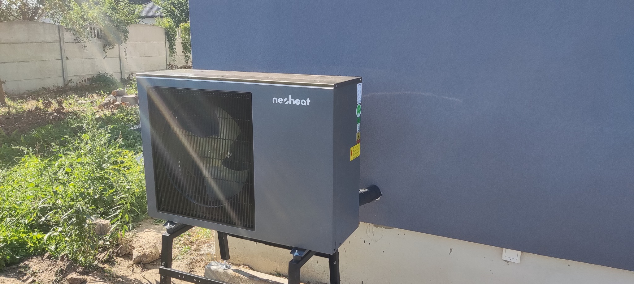 Szara pompa ciepła marki Neoheat zamontowana na zewnątrz budynku, na metalowej konstrukcji, z widocznymi elementami instalacji.