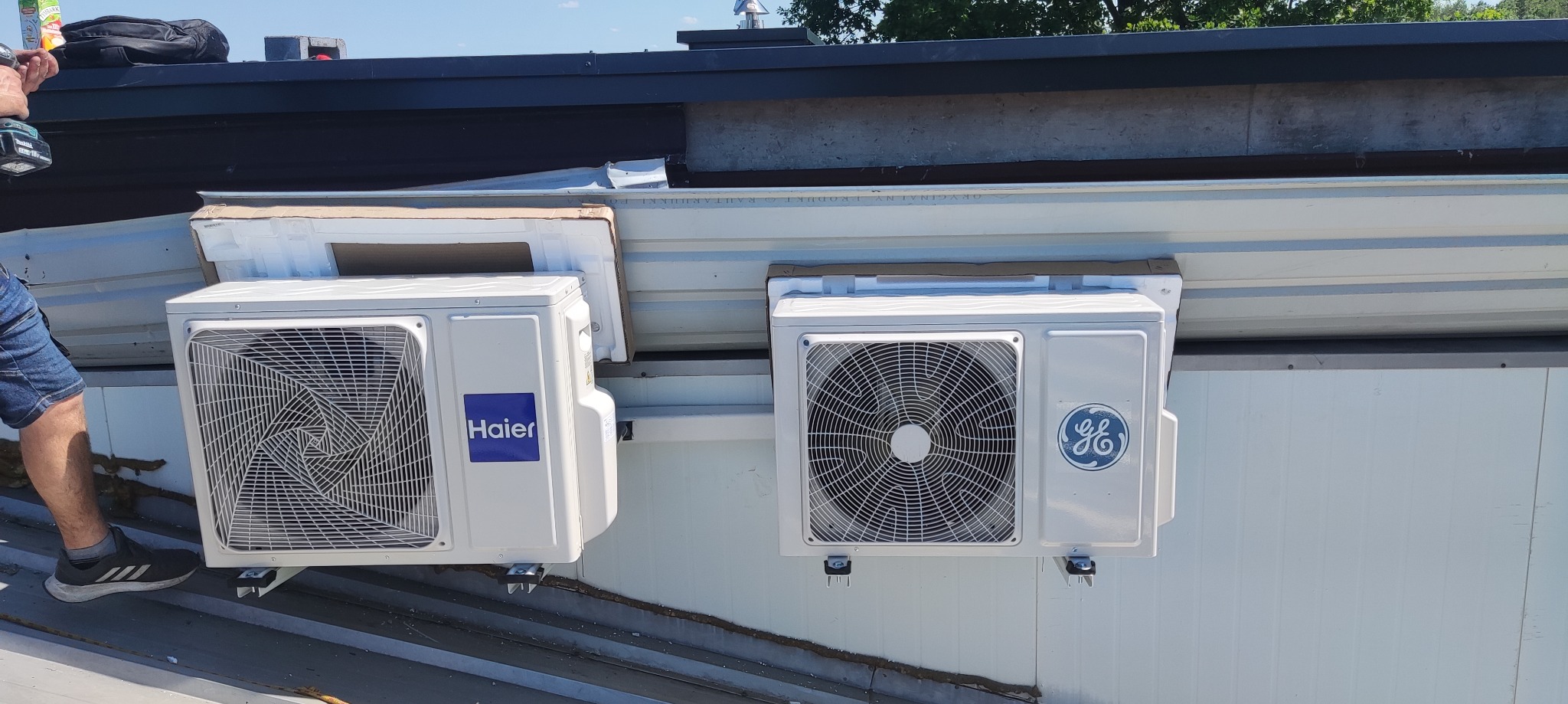 Dwa zamontowane klimatyzatory różnych marek (Haier i GE) na zewnątrz budynku, widoczne elementy instalacji i fragment osoby montującej.