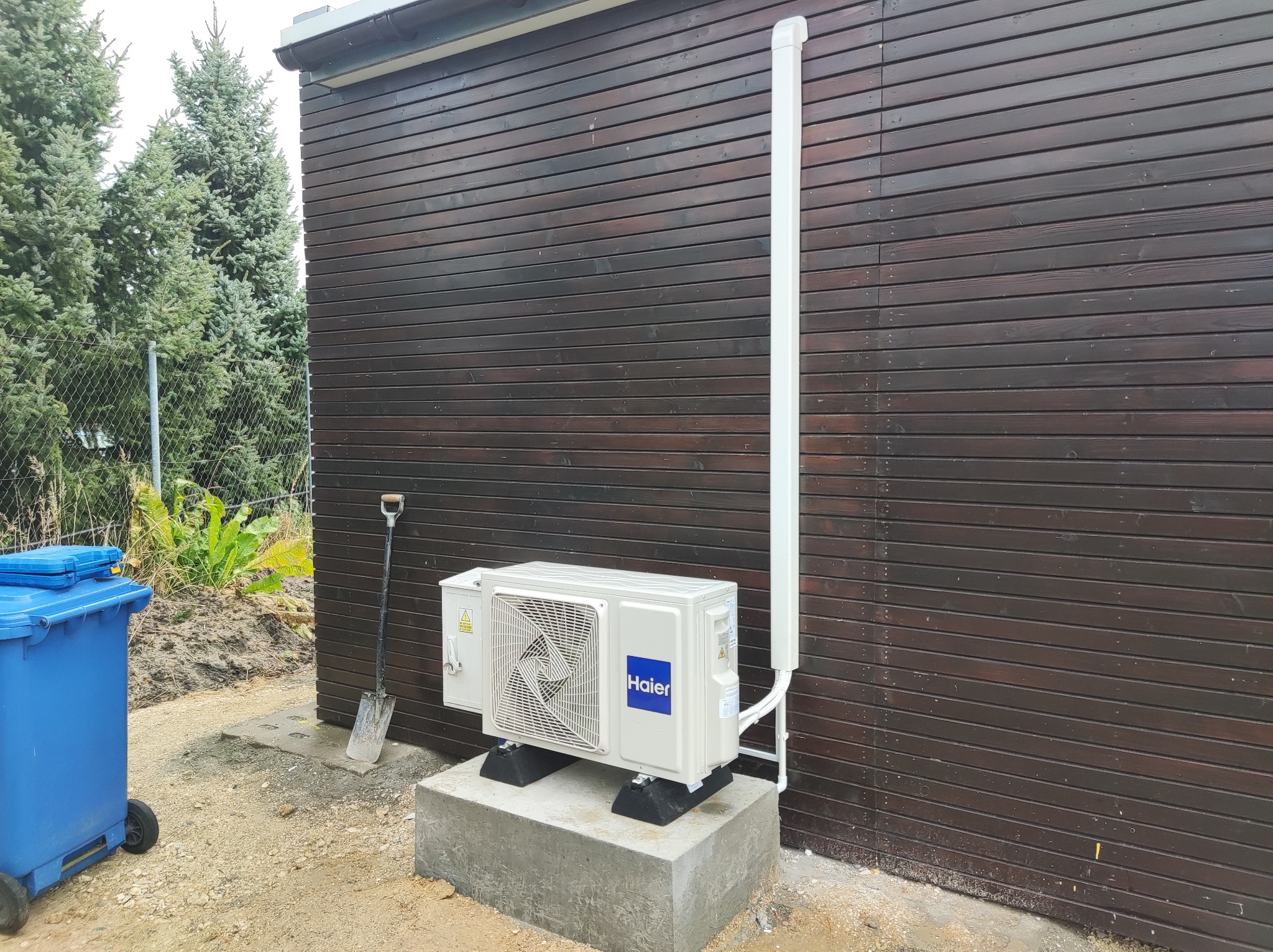 Zewnętrzna jednostka klimatyzacji Haier zamontowana na betonowym fundamencie przy drewnianej ścianie budynku. Obok stoi niebieski kosz i oparta jest łopata.