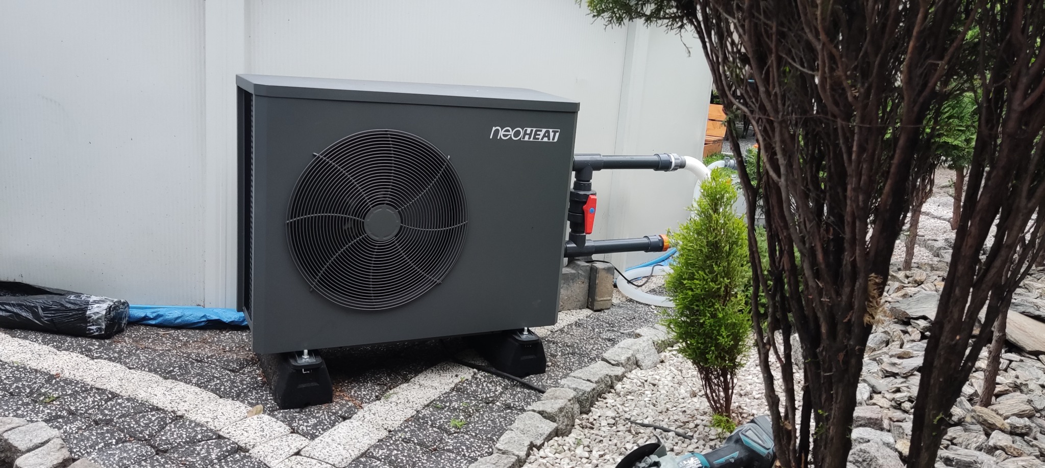 Szara pompa ciepła Neoheat na zewnątrz budynku, z widocznymi rurami i elementami instalacji, otoczona zielenią i kamieniami. Widoczny fragment narzędzia.