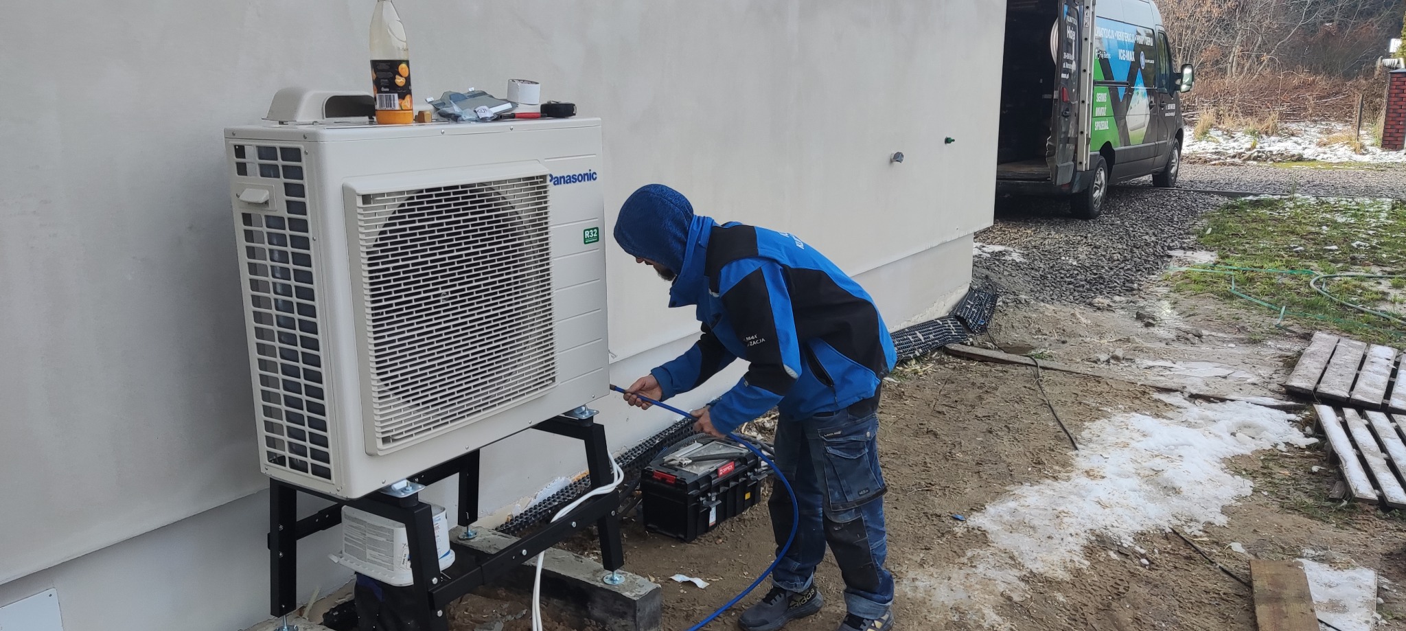 Instalator w trakcie montażu zewnętrznej jednostki klimatyzacji Panasonic na metalowej konstrukcji przy ścianie budynku. Widoczny samochód serwisowy w tle.