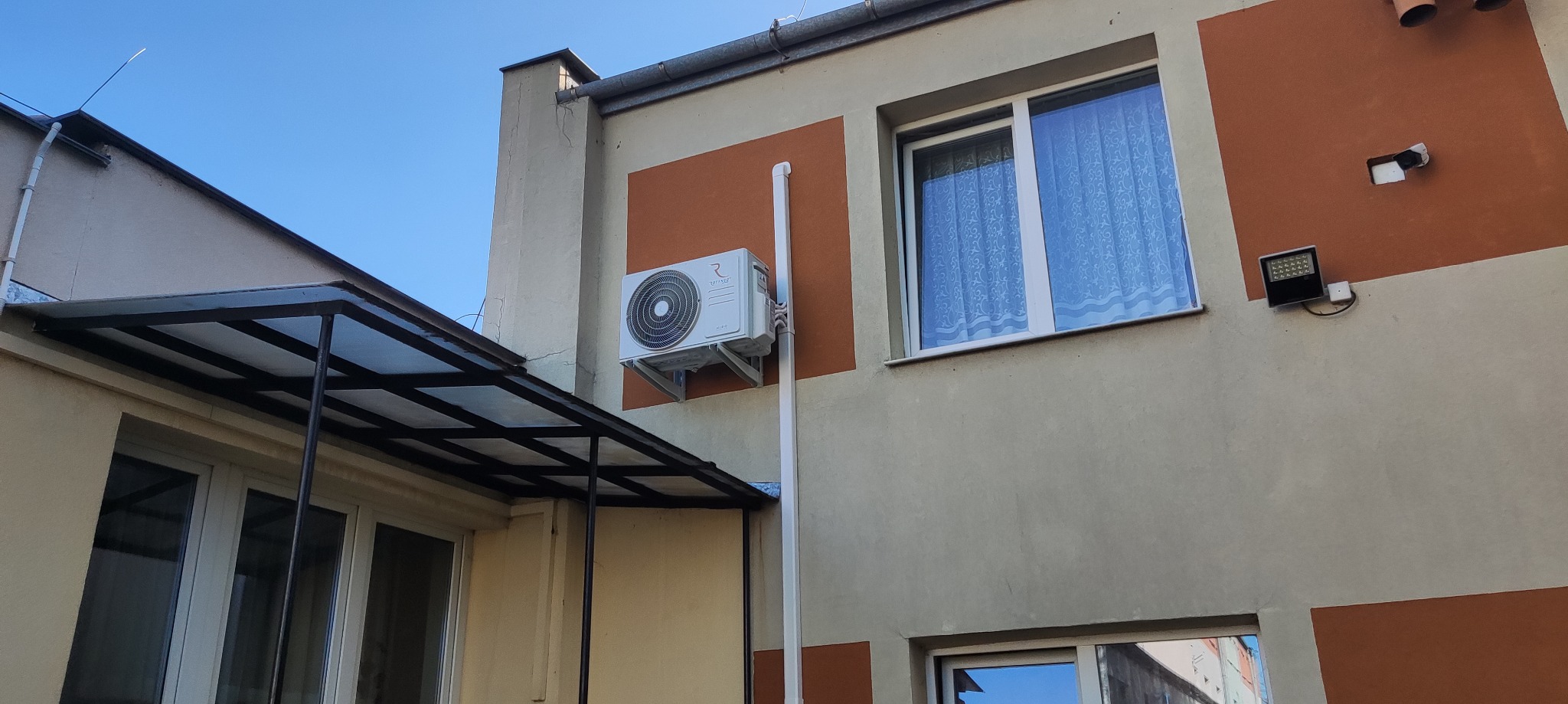 Biały klimatyzator zamontowany na elewacji budynku z beżowo-pomarańczowymi akcentami, widoczny z zewnątrz, z rurą odprowadzającą. Nad nim okno z firanką.