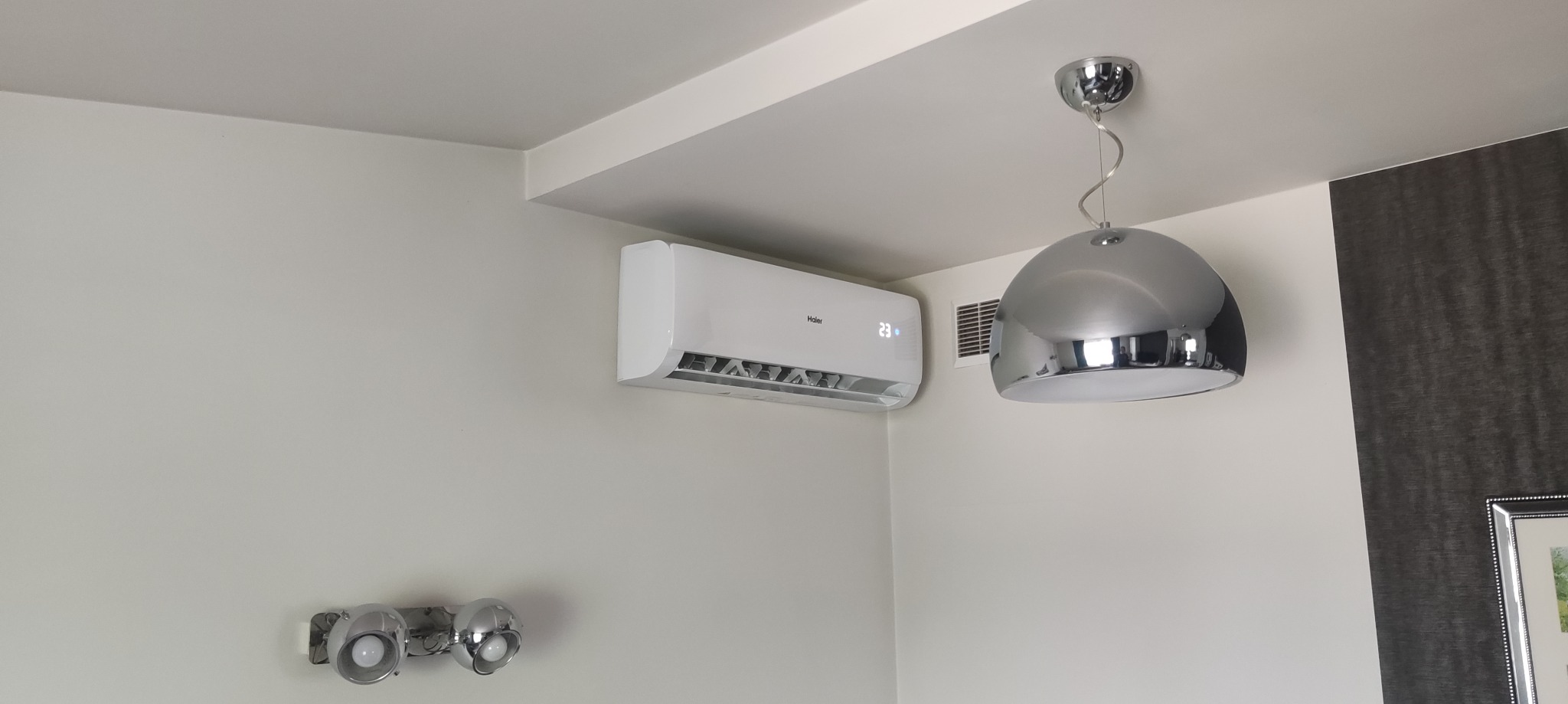 Biały klimatyzator Haier na ścianie w jasnym wnętrzu, obok designerska lampa wisząca i oświetlenie punktowe. Widoczny wyświetlacz temperatury.