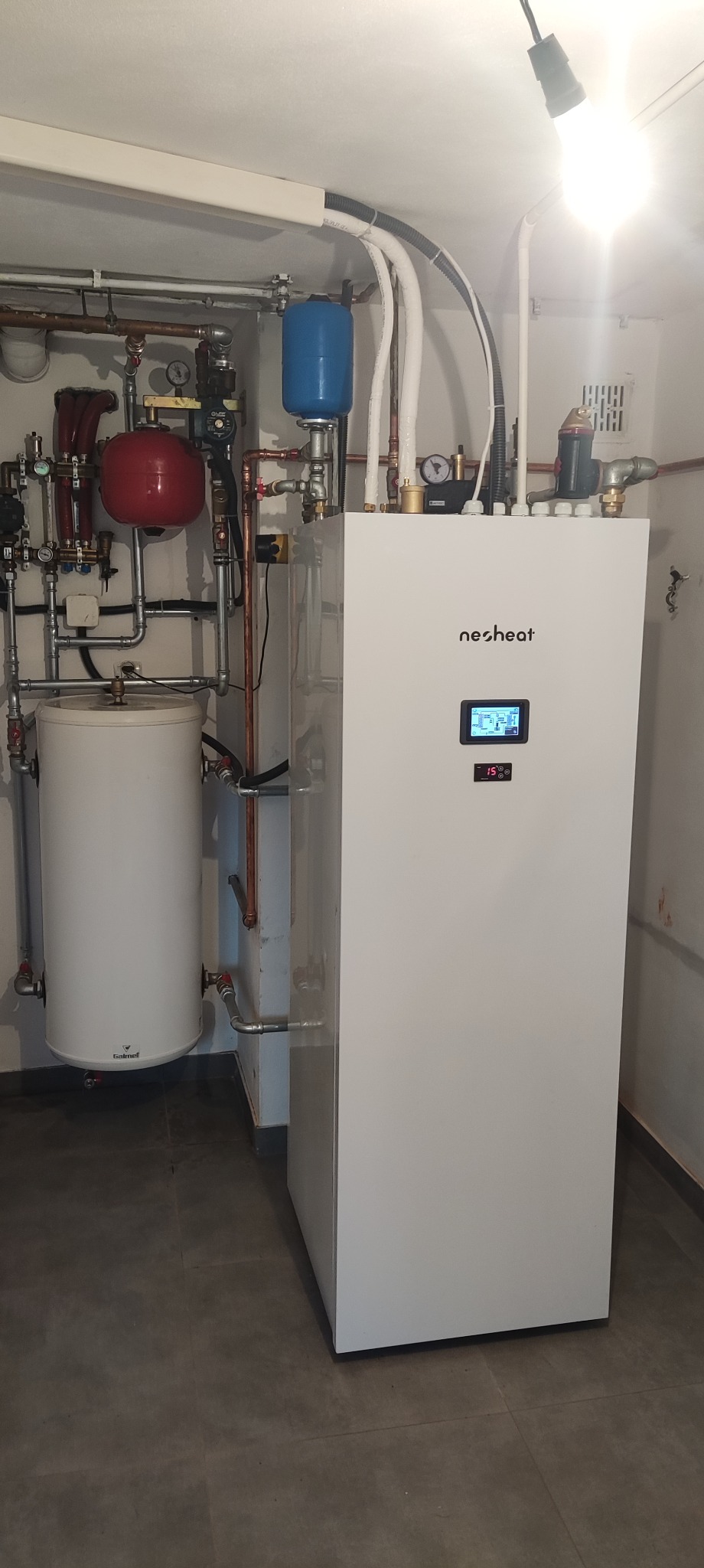 Biała pompa ciepła marki Nesheat z podłączonymi instalacjami, zbiornikami i rurami w kotłowni. Widoczny wyświetlacz z temperaturą 15 stopni Celsjusza.