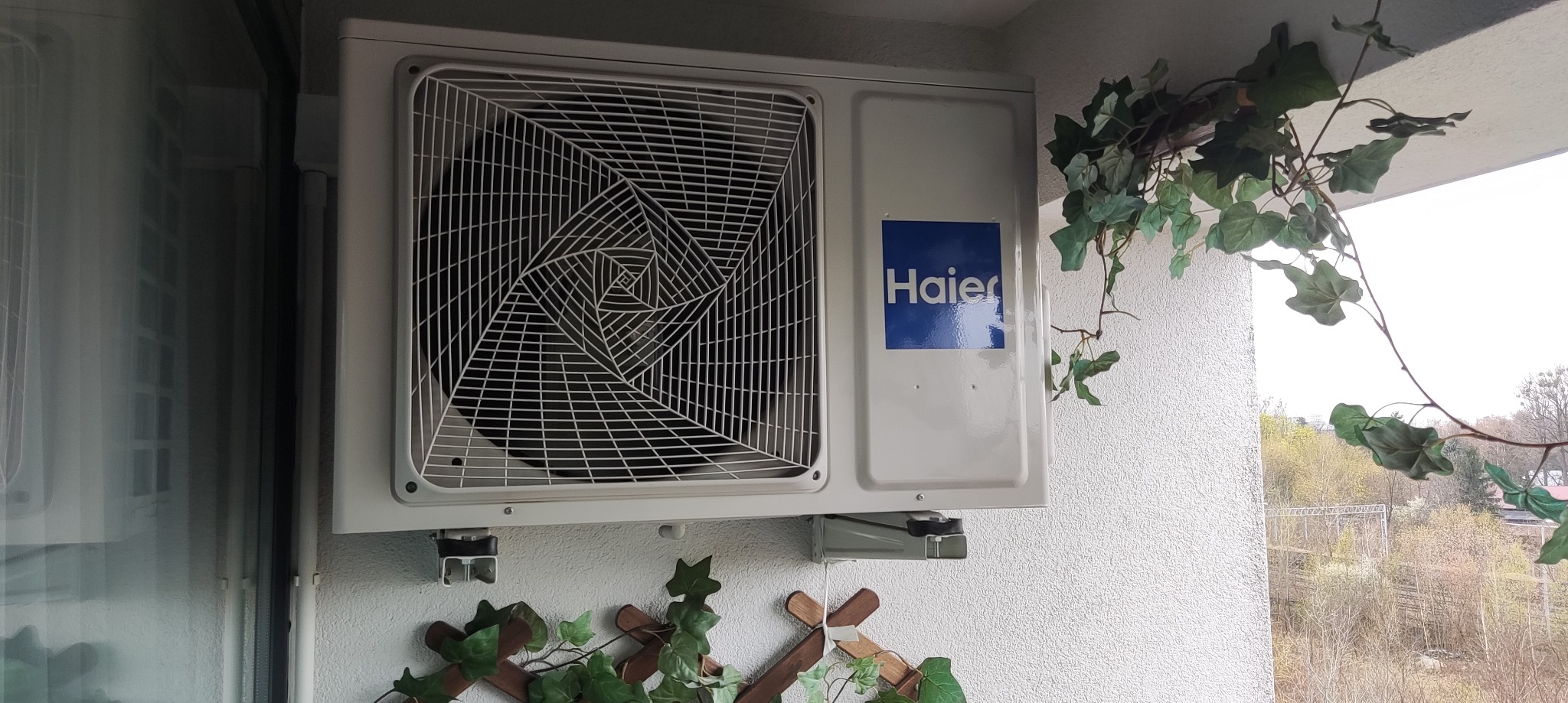 Biała jednostka zewnętrzna klimatyzacji Haier zamontowana na balkonie, z dekoracyjną zielenią. Widok na zewnątrz przez balustradę.