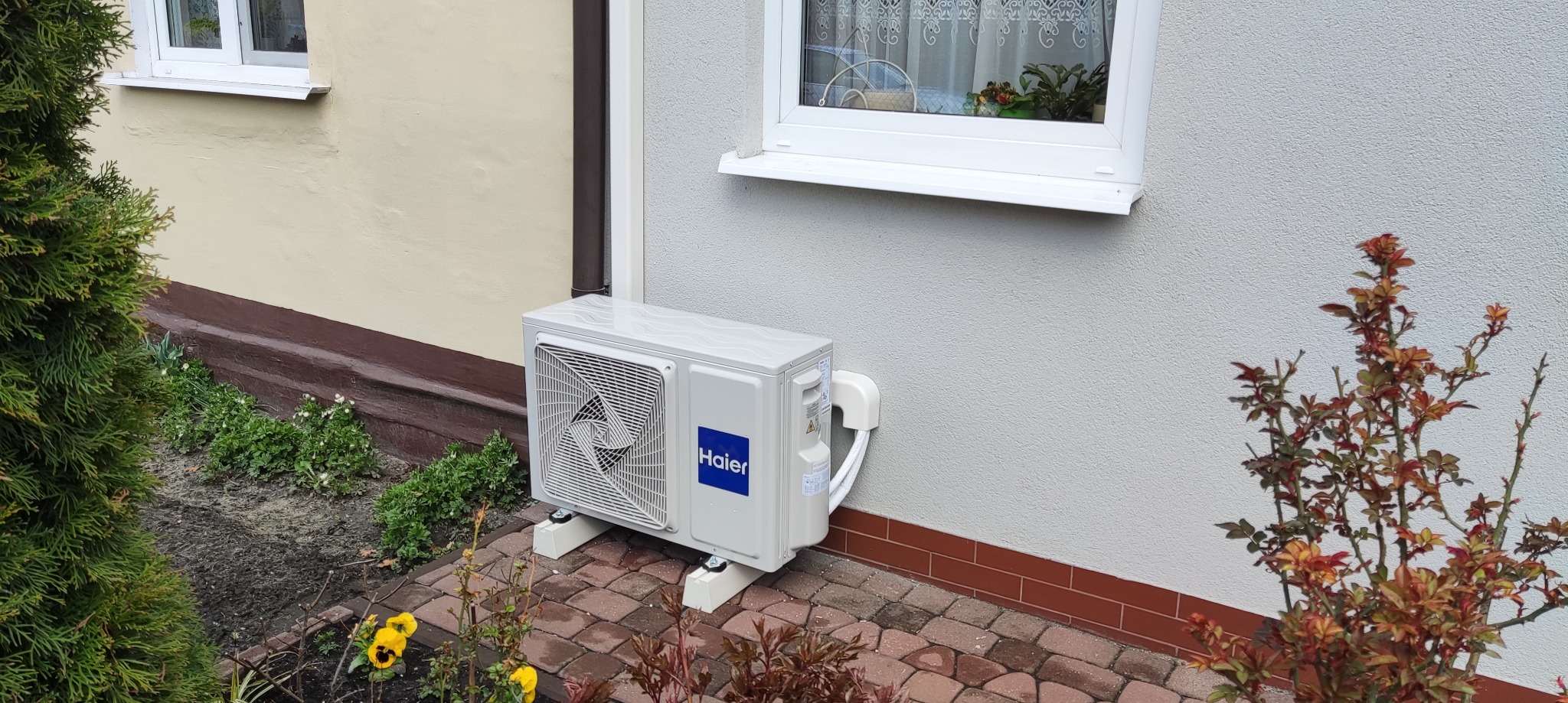 Zewnętrzna jednostka klimatyzacji Haier zamontowana na kostce brukowej przy ścianie domu z oknem. Widoczne rośliny w tle i rynna spustowa.