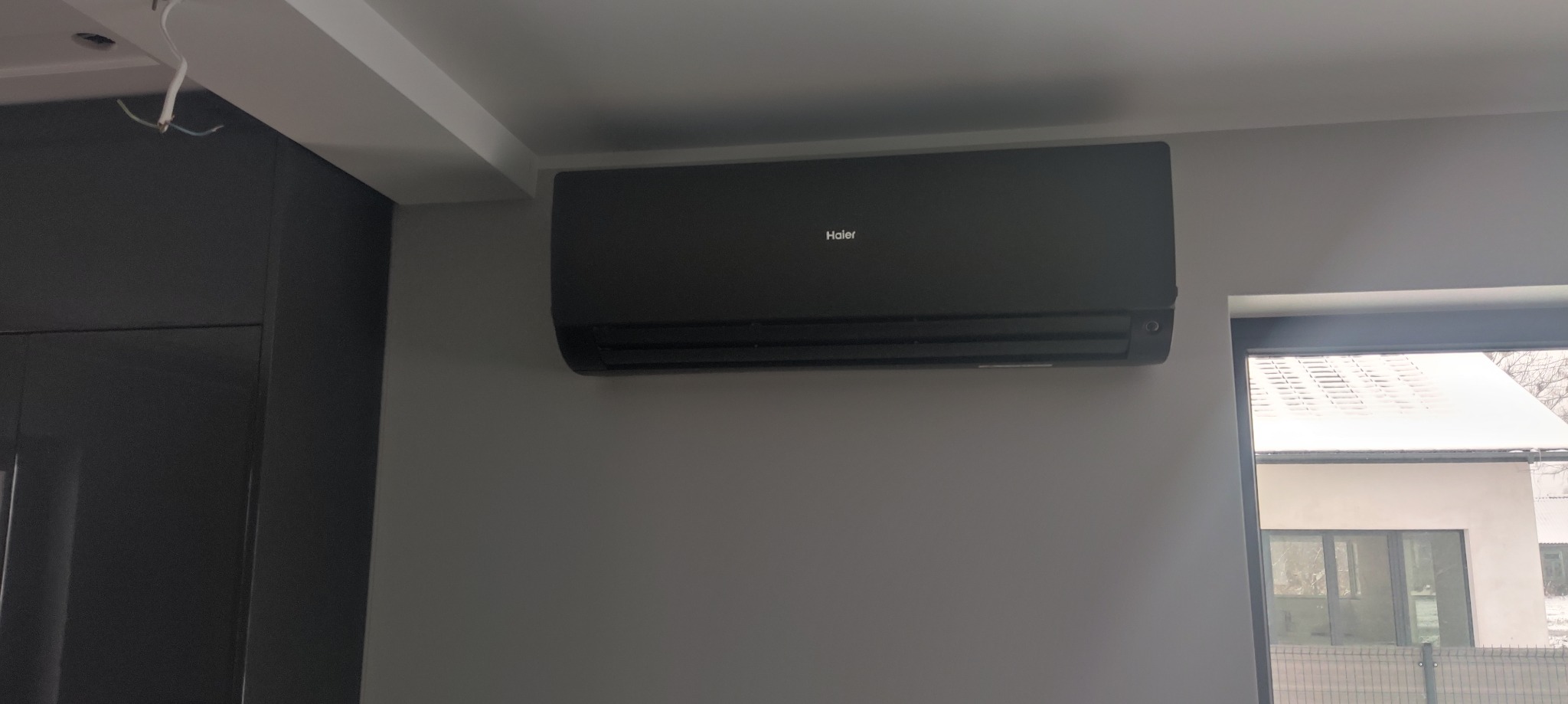 Czarny klimatyzator Haier zamontowany na szarej ścianie w nowoczesnym wnętrzu, widok z okna na zewnątrz. Minimalistyczny design.
