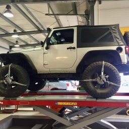 Auto Service Domagała - Niezależny Warsztat Ford - Biały Jeep Wrangler na podnośniku w warsztacie, z zamontowanymi urządzeniami do geometrii kół. Widoczne opony terenowe i elementy wyposażenia warsztatowego.