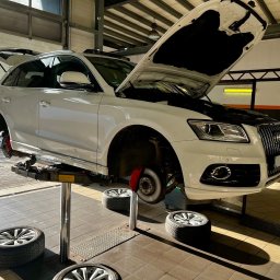 Auto Service Domagała - Niezależny Warsztat Ford - Białe Audi Q5 na podnośniku w warsztacie, z otwartą maską i zdjętymi kołami, ukazujące tarczę hamulcową. W tle brama garażowa i oświetlenie warsztatowe.