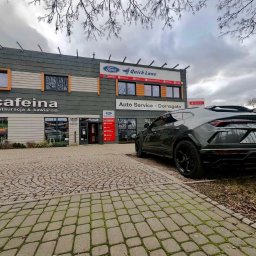 Auto Service Domagała - Niezależny Warsztat Ford - Warsztat samochodowy Ford Quick Lane z zaparkowanym Lamborghini Urus. Widoczny szyld Auto Service Domagała i kawiarnia Cafeina. Brukowany podjazd, pochmurne niebo.