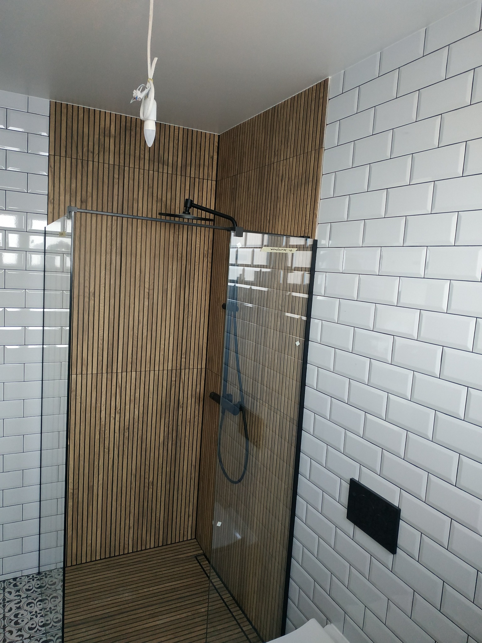 Nowoczesna kabina prysznicowa z drewnianymi panelami i białymi płytkami typu metro. Minimalistyczny design, czarna armatura, widoczne detale wykończenia.