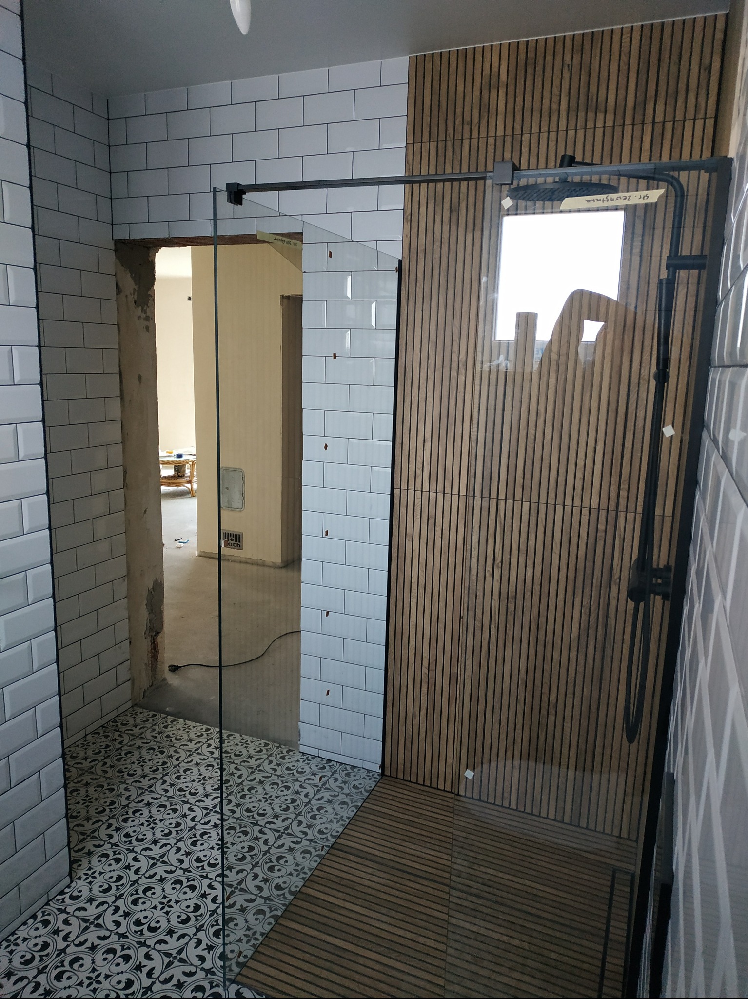 Nowoczesna łazienka po remoncie: białe płytki typu metro, drewniane panele ścienne, wzorzysta podłoga i czarna armatura prysznicowa. Widok na sąsiednie pomieszczenie.