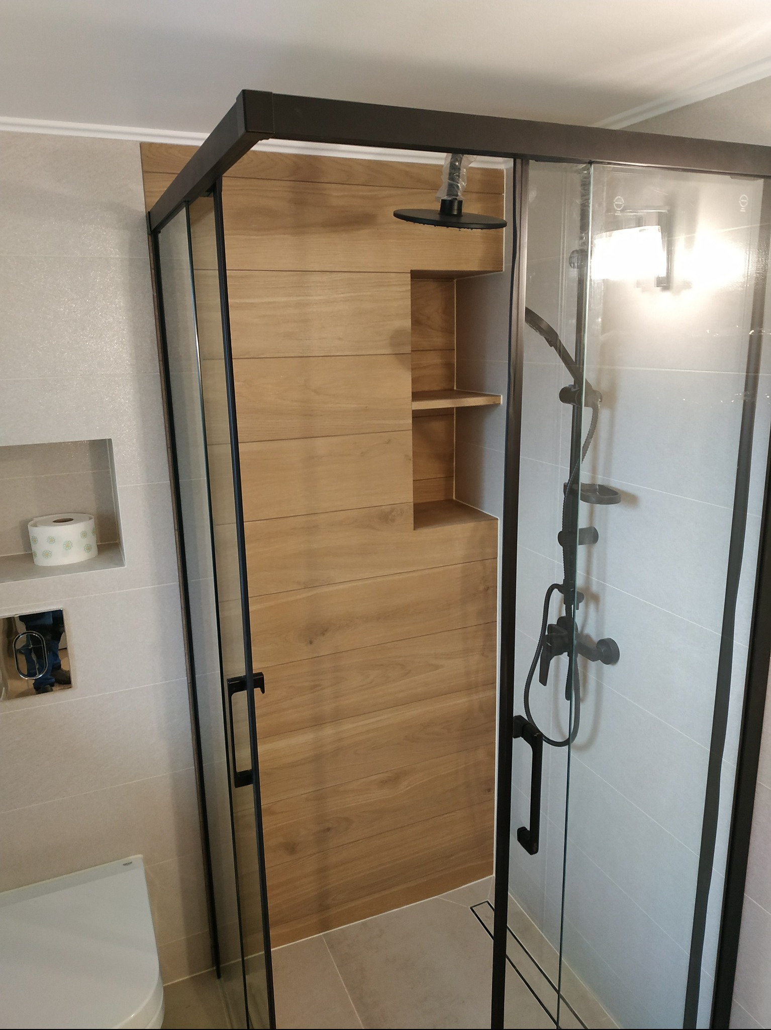 Nowoczesna kabina prysznicowa z czarną ramą i deszczownicą, wykończona płytkami imitującymi drewno. Widoczna toaleta i wnęka z papierem toaletowym.
