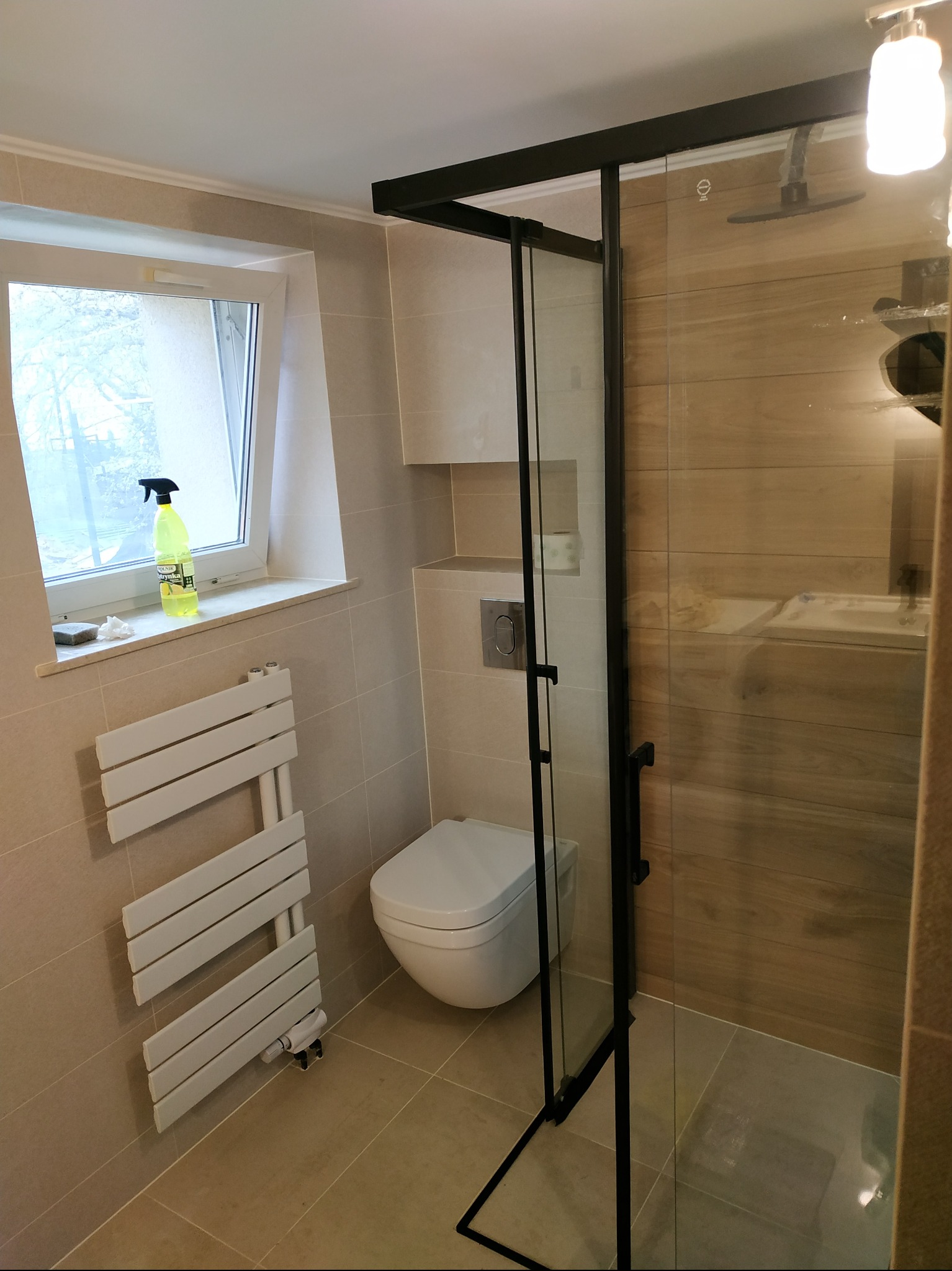 Jasna łazienka po remoncie: beżowe płytki, kabina prysznicowa z czarną ramą, toaleta, grzejnik drabinkowy i okno z płynem do czyszczenia.
