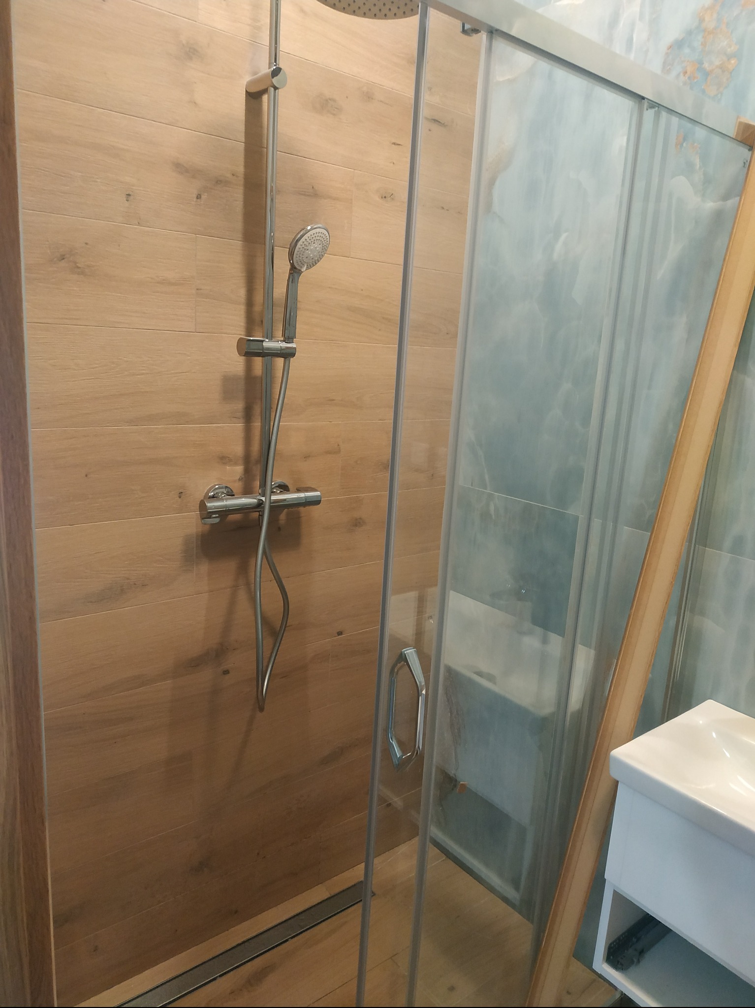 Kabina prysznicowa z deszczownicą i panelami imitującymi drewno, widok na umywalkę i niebieskie płytki. Nowoczesna aranżacja łazienki po remoncie.