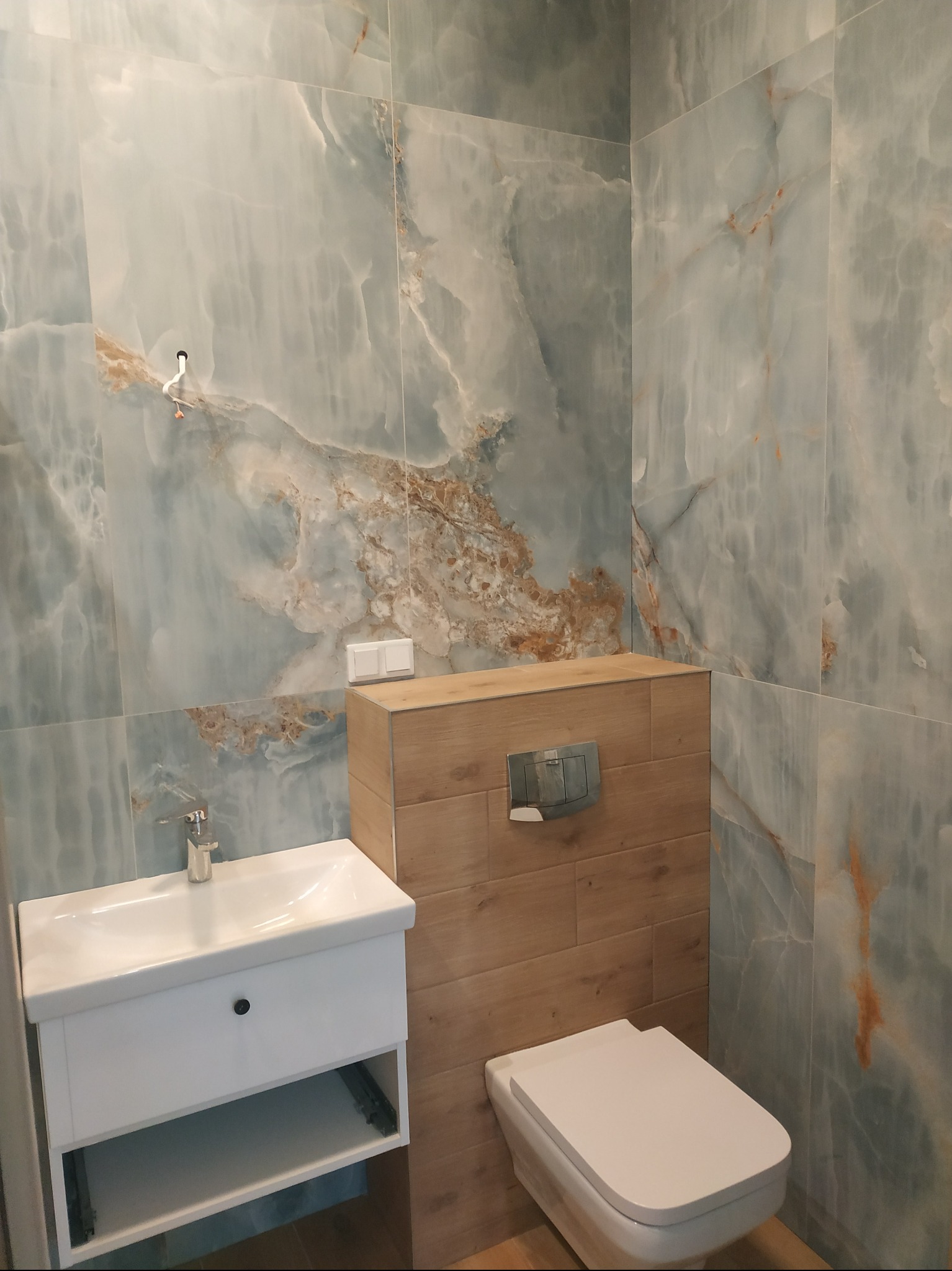 Wykończona łazienka w Opole: marmurowe płytki w odcieniach błękitu i brązu, podwieszana toaleta z drewnianą obudową i biała umywalka z szafką.