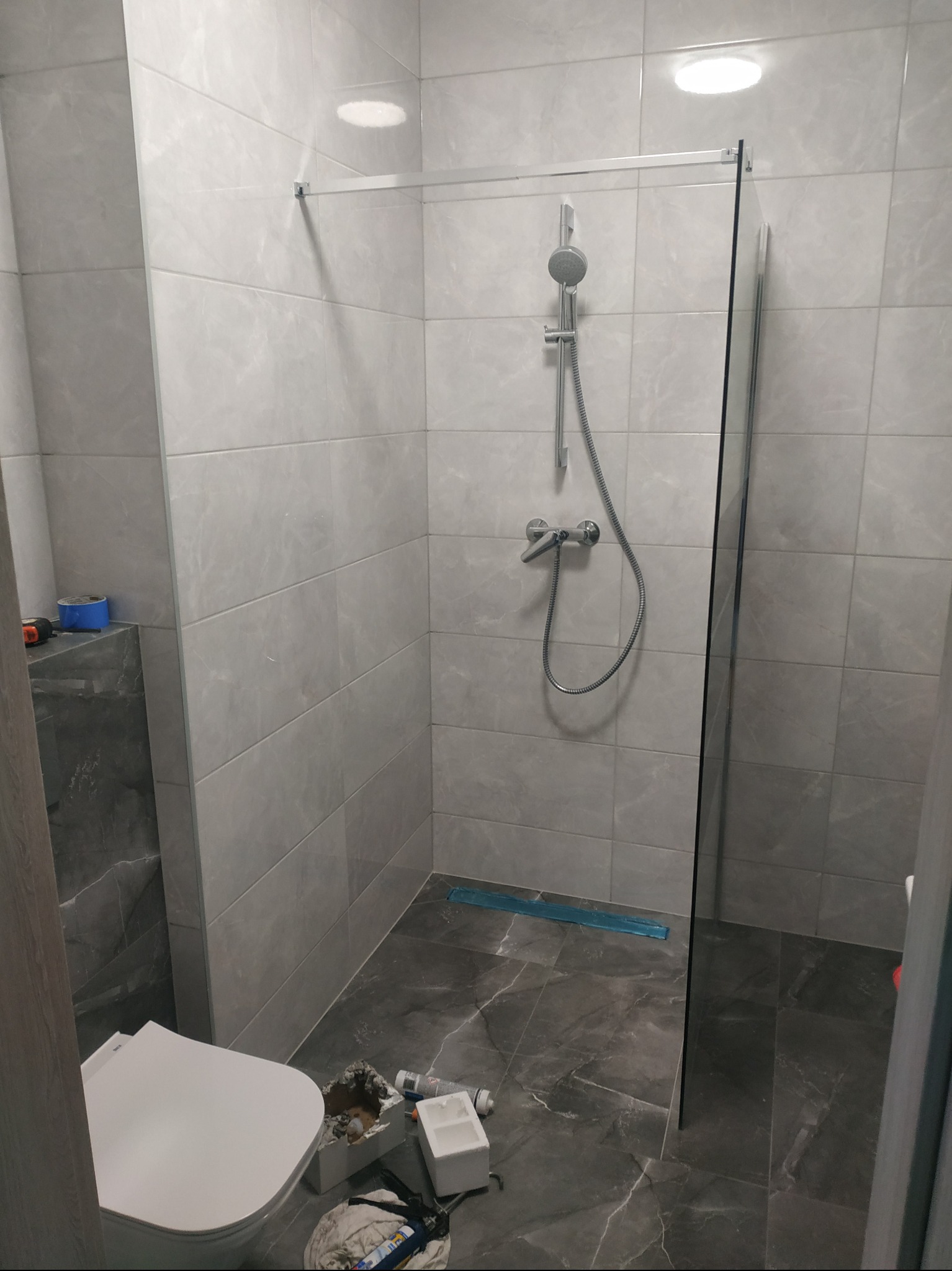 Nowoczesna kabina prysznicowa z szarymi płytkami i szklaną ścianką. Widoczny odpływ liniowy i częściowo zamontowana toaleta. Użyte materiały budowlane na podłodze.